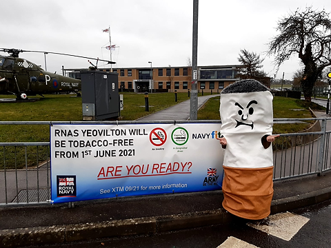 RNAS Yeovilton tobacco free banner on railing, with person in a stubbed cigarette mascot costume standing on the right side