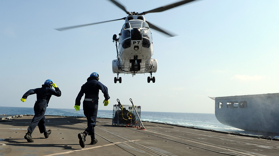 Royal Navy personnel approaching a landing helicopter on the flight deck. 