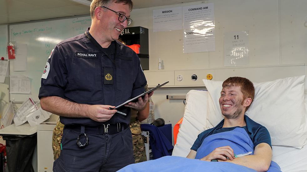 male naval nurse holding clipboard speaking to patient in bed