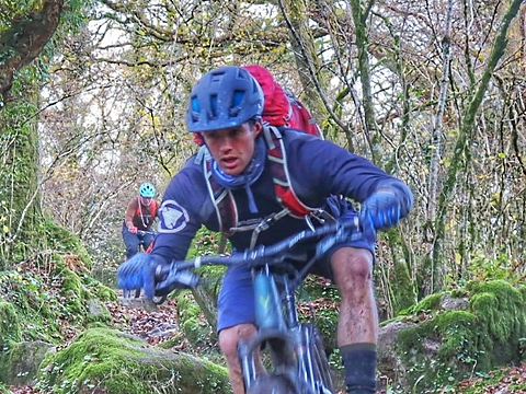 close up of mountain biker tackling course through wooded trail, with competitor following close behind