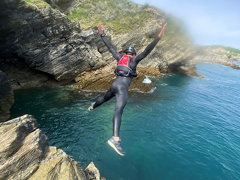 coasteerer dressed in black wetsuit and helmet  leaping off cliff face into blue sea