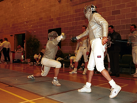 two fencers in indoor event wit others watching, one fencer on one knee defending, other standing on attack