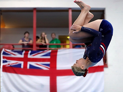 gymnast in mid-air, completing tumble as part of floor routine with rn white ensign and small gallery of spectators in background