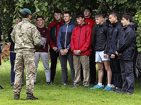 orienteering group in grassed area and under a tree,  being briefed by fatigued-dressed instructor
