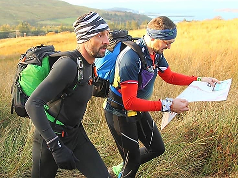 two orienteers laden with backpacks crossing an uncropped hay-filled meadow, one, dressed in black trousers and red and blue top, consulting a map the other, dressed all in black, focused on the way ahead
