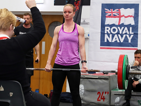 weightlifter in pick top, black joggers being given instruction in technique, with royal navy ensign logo on wall behind