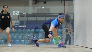 royal navy male squash player in blue stooping low for backhand with black dressed female opponent watching on to the left