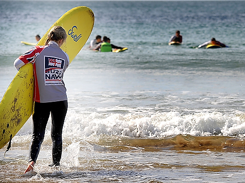 surfer in grey, blue, white, red rn kit entering water, carrying yellow board