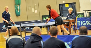 male table tennis player in red reaching for ball after hitting it into the net against blue dressed female opponent