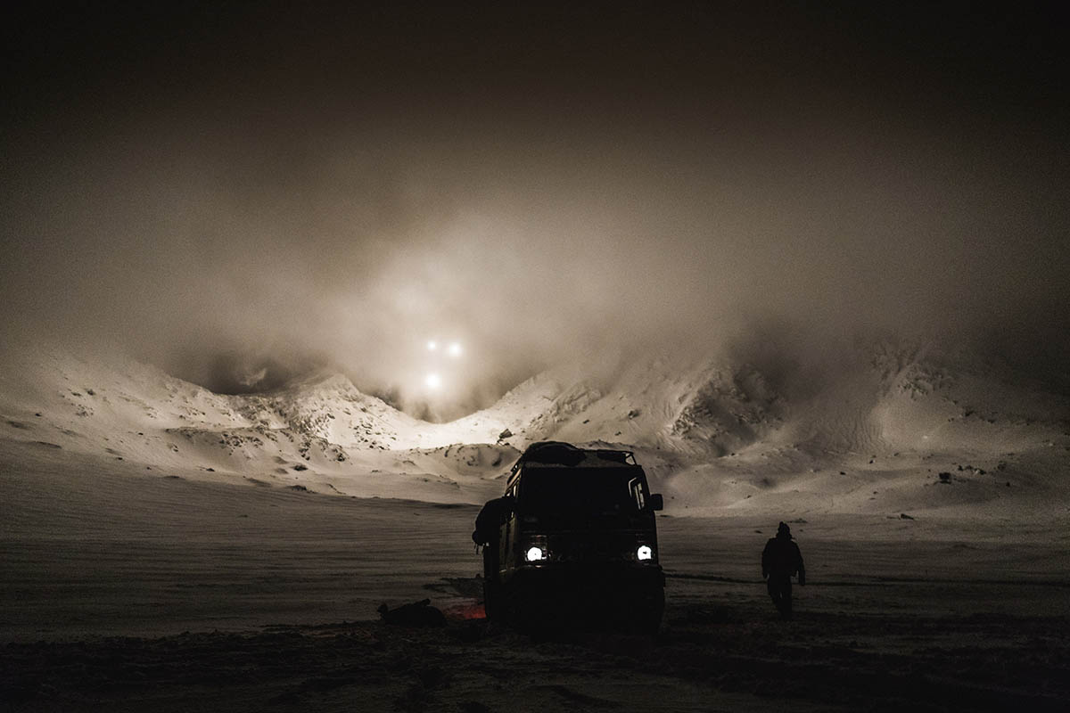A BV all-terrain vehicle and a Royal Marine silhouetted against a Norwegian mountain range is lit up at night by mortars illumination rounds